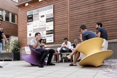 Bürogebäude aus Holz von außen mit Tafel von Firmenlogos. Davor sitzen Personen und essen Eis.
