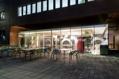 Cafeteria aus Glas, beleuchtet von Außen, im Vordergrund Tische und Stühle im Freien