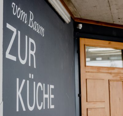 Links schwarze Wand mit der Aufschrift "vom Baum ZUR KÜCHE", rechts Holztüre