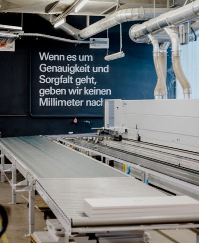 Sägemaschine und Fließband, hinten Wand mit Aufschrift " Wenn es um Genauigkeit und Sorgfalt geht, geben wir keinen Milimeter nach."