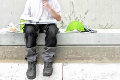 Bauarbeiter, der Pause macht. Den Helm hat er abgelegt - auf seinem Schoß liegt eine Mappe, in der er liest. Im Bild außerdem: Wurstsemmel und Getränkeflasche.