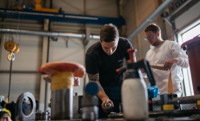 Ein Arbeiter in Schwarz sprüht mit Spraydose, dahinter Arbeiter in weißem Schutzanzug