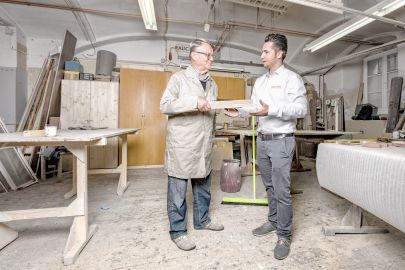 © Niki Schreinlechner Älterer Mann übergibt jüngerem Mann in Werkstatt ein Staffelholz.