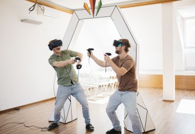 Zwei Männer mit Virtual-Reality-Brillen und Joysticks in der Hand vor weißem Bogen aus Papier.