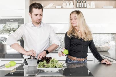 © Niki Schreinlechner Ein Mann und eine Frau bereiten Salat zu. Der Mann presst eine Limette mit einer herkömmlichen Zitruspresse aus; die Frau hat eine Sprühvorrichtung in eine Limette gesteckt und zeigt dem Mann, wie es auf diese Weise einfacher geht.