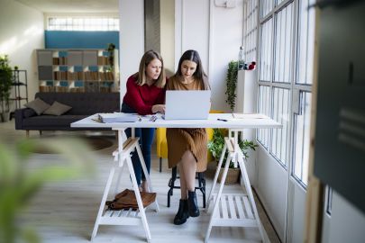 (c) Westend61_Giorgio_Fochesato Geschäftsfrau lehnt sich an eine Kollegin, die einen Laptop am Schreibtisch im Büro benutzt.